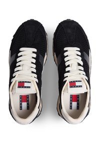 Μαύρα αθλητικά παπούτσια από διχτυωτό υλικό και σουέντ με μπεζ κορδόνια, γκρι λεπτομέρειες και γλώσσα με λογότυπο που φέρει κόκκινες, λευκές και μπλε ρίγες.