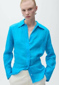 Camicia turchese a maniche lunghe con colletto a punta e chiusura con bottoni. Realizzata in un tessuto leggero, presenta una vestibilità rilassata e una consistenza liscia.