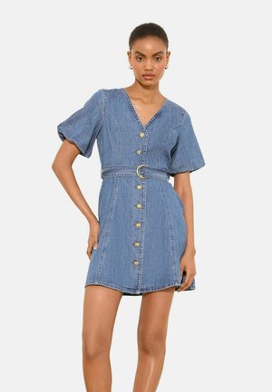 Naine kannab sinist sametist nööbitustega kleiti puffy lühikeste varrukatega ja vöökohal vööga, selge heleda tausta ees.