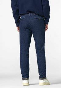 Jean en denim bleu foncé avec une coupe ajustée, comportant une taille avec des détails subtils, deux poches arrière et une finition soignée aux chevilles.