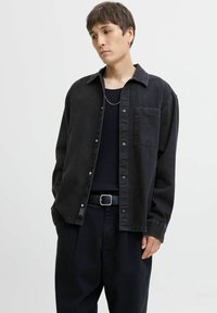 Black denim overshirt with snap buttons, single chest pocket, and structured fit, paired with a ribbed black top and dark trousers.