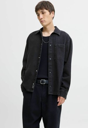 Sort denim overshirt med trykknapper, enkelt brystlomme og struktureret pasform, parret med en ribbet sort top og mørke bukser.