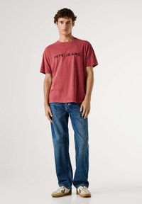 Camiseta roja de algodón con texto negro "PEPE JEANS", mangas cortas y cuello redondo, combinada con vaqueros azul rectos y zapatillas blancas.