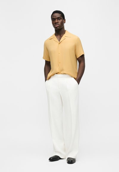 Άνδρας που φοράει μουσταρδί κοντομάνικο πουκάμισο με κουμπιά, λευκό παντελόνι φαρδύ στο κάτω μέρος και μαύρα παπούτσια, όρθιος με τα χέρια στις τσέπες.