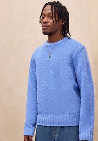 Νεαρός άνδρας με πλεξούδες που φοράει ένα ανοιχτό μπλε χνουδωτό πουλόβερ και κρεμαστό κολιέ, στέκεται μπροστά σε μια απλή μπεζ φόντο.