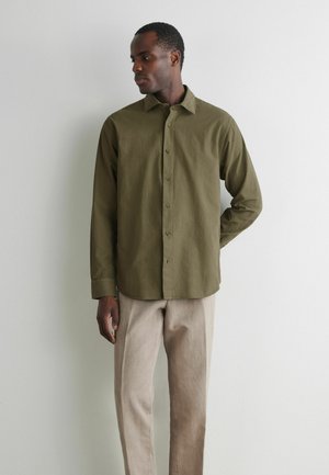 Mann steht an einer weißen Wand, trägt ein olivgrünes Hemd mit Knöpfen und beige Hosen, blickt nach links und hat eine Hand hinter dem Rücken.