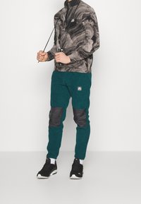 Heren zip-up jas met zwart en grijs gemarmerd patroon; teal fleece broek met zwarte kniestukken; zwarte sneakers.