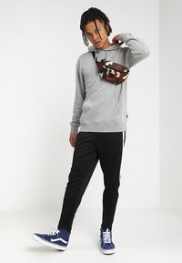 Felpa grigia, pantaloni sportivi neri, sneakers blu navy e una borsa a tracolla a camo multicolore indossata sul petto. L'ambientazione è neutra.