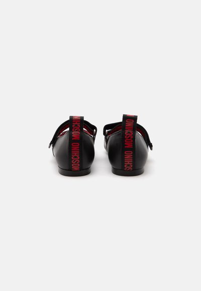 Paire de chaussures plates noires brillantes avec le texte rouge « Moschino » sur les languettes arrière du talon, vues de dos sur fond blanc.