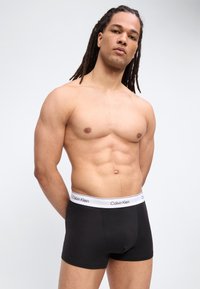 Paita pois päältä pitkäkiharainen mies mustissa Calvin Kleinin bokserialushousuissa seisoo yksinkertaisen vaalean taustan edessä.