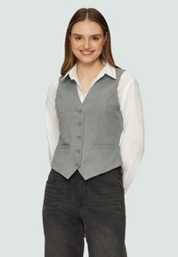 Grijze fitted vest van stof, met een V-hals, zes knopen en twee voorzakken, gecombineerd met een witte overhemd met kraag en donkere broek.