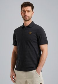 Zwarte polo met een kraag en drie knopen, met een gestructureerd patroon en een gouden logo op de linkerkant van de borst. Korte mouwen.