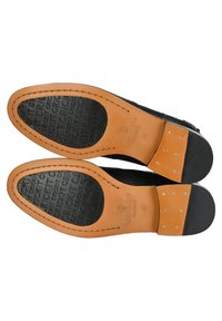 Suole di scarpe in pelle nera con accenti marroni. Forma arrotondata, grip in gomma nera testurizzata, cuciture visibili e marchio.