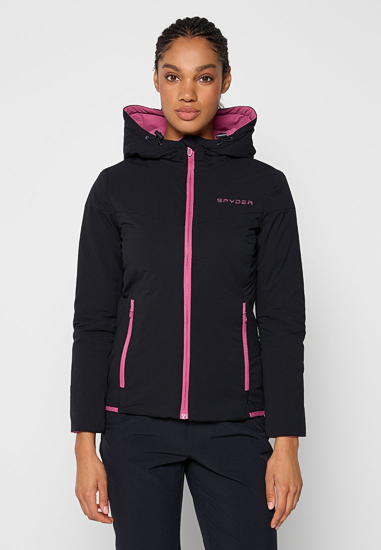 Femme portant une veste à capuche noire avec une fermeture éclair rose, des bordures de poche roses et le logo "SPYDER" sur la poitrine, debout devant un fond uni.