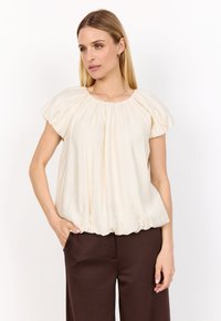 Blus med puffärmar i krämfärgat tyg, med en draperad halsringning, mjukt strukturerad textil och rundad nederkant, kombinerad med brun högmidjade byxor.