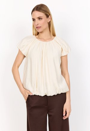 Blus med puffärmar i krämfärgat tyg, med en draperad halsringning, mjukt strukturerad textil och rundad nederkant, kombinerad med brun högmidjade byxor.