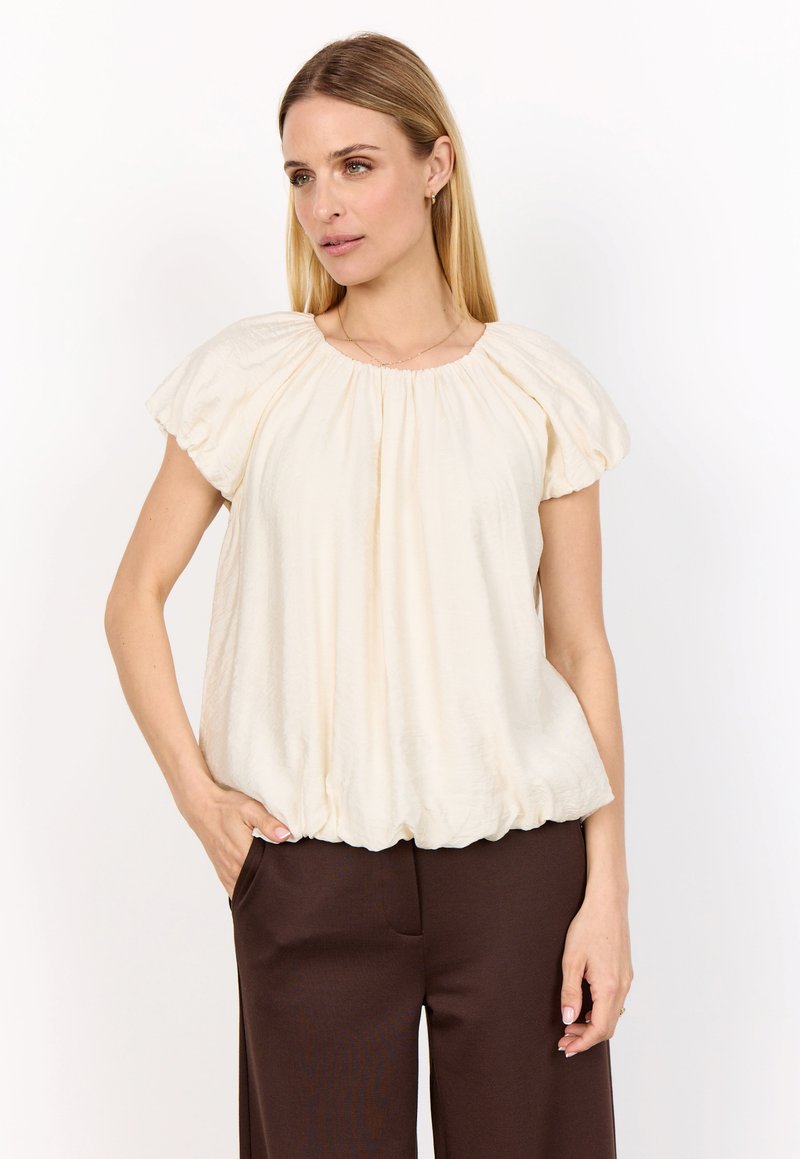 Blus med puffärmar i krämfärgat tyg, med en draperad halsringning, mjukt strukturerad textil och rundad nederkant, kombinerad med brun högmidjade byxor.