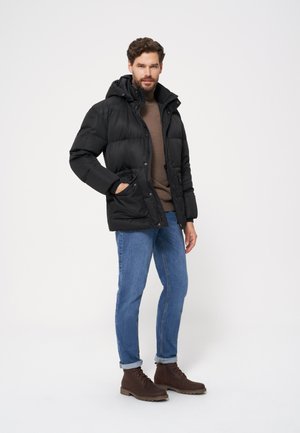 Czarna kurtka puchowa z kapturem, zapinana na zamek, z dwiema bocznymi kieszeniami, noszona na brązowym swetrze, w połączeniu z niebieskimi dżinsami i brązowymi botkami.