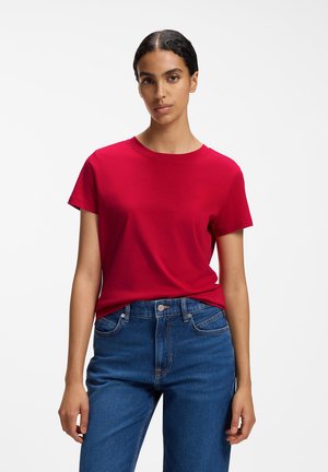 Frau mit dunklen Haaren, die ein schlichtes rotes T-Shirt mit kurzen Ärmeln und blaue Jeans trägt, steht vor weißem Hintergrund.