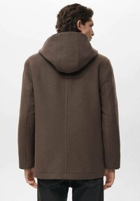 Hombre con abrigo con capucha marrón de espaldas, mostrando la vista trasera, vistiendo pantalones oscuros contra un fondo claro y liso.