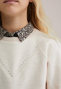 Šviesiaplaukis asmuo dėvi smėlio spalvos megztinį su sidabriniais užtrauktukais ir leopardų rašto marškinių apykaklę, stovintis prieš paprastą foną.