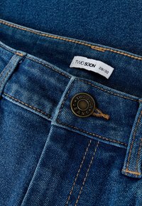 Mėlynų džinsų juosmens artimas vaizdas, kuriame matomas metalinis mygtukas ir baltas etiketė su prekės ženklu „Two Soon“ bei dydžiu 28/32.