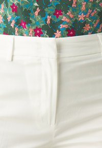 Pantalones blancos ajustados con una textura suave y bolsillos frontales, combinados con una blusa verde con estampado que presenta flores y hojas rosas.