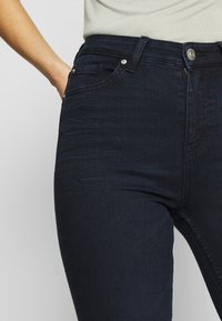 Jeans de mezclilla azul oscuro con corte slim, tela texturizada, bolsillos frontales y herrajes metálicos en la cintura.