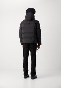 Czarna kurtka puchowa z kapturem, o poziomym pikowanym wzorze, błyszczącej teksturze, noszona z czarnymi spodniami i butami, widok z tyłu.