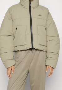 Cropped puffer jacket in light beige fabric, featuring a high collar, black zip closure, and two front pockets. Textured surface throughout.