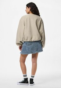 Vrouw staat met de rug naar ons toe en draagt een beige sweatshirt, een blauwe denim mini-jurk, gestreepte witte sokken en zwarte high-top sneakers.