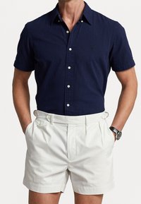 Navy short-sleeve shirt with textured fabric and button-down collar, paired with white pleated shorts featuring side pockets and adjustable hardware.
