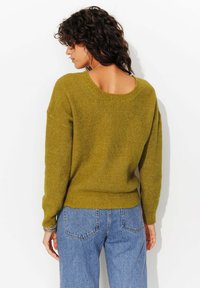 Pull en tricot vert olive avec un col large et des épaules tombantes, associé à un jean en denim bleu clair. Design texturé et ample.