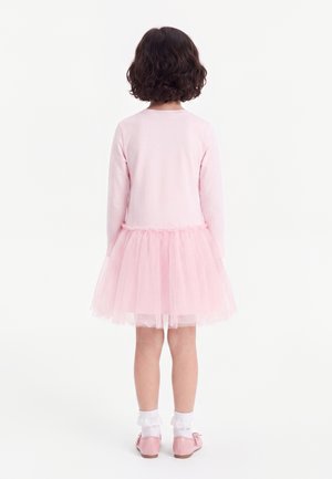 Enfant aux cheveux courts et bouclés portant un haut à manches longues rose clair, une jupe en tulle rose, des chaussettes blanches à volants et des chaussures roses, debout de dos.