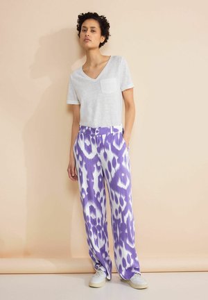 T-shirt blanc à col en V avec une poche, associé à un pantalon ample à motif violet et blanc. Le mannequin porte des baskets blanches. Fond simple.