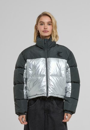 Padded jacket with shiny silver front and matte dark green sleeves, high collar, and zip closure. Features a logo on the chest.