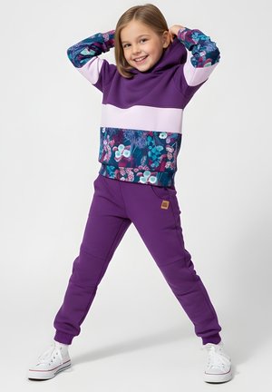 Uśmiechnięta dziewczyna pozująca z rękami za głową, ubrana w fioletową bluzę z kwiatowym wzorem i pasujące spodnie oraz białe trampki na białym tle.