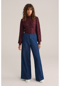 Bordo spalvos ilgomis rankovėmis palaidinė su iškilusiu gėlių raštu, derinama su tamsiai mėlynais, aukštu liemeniu ir plačiomis kojų kelnėmis iš džinso. Modelis stovi priešais neutralią fono sieną.