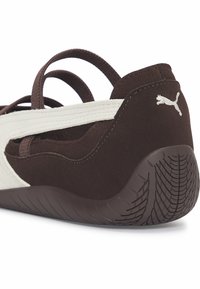 Καφέ αθλητικό παπούτσι από σουέντ με λευκή πλευρική ρίγα, διαθέτει διασταυρωμένους ιμάντες και καφέ σόλα από καουτσούκ με υφή. Εμφανίζεται το λογότυπο Puma.