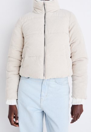 Personne portant une doudoune côtelée beige avec une fermeture éclair à l'avant et un jean taille haute bleu clair, debout sur un fond blanc.