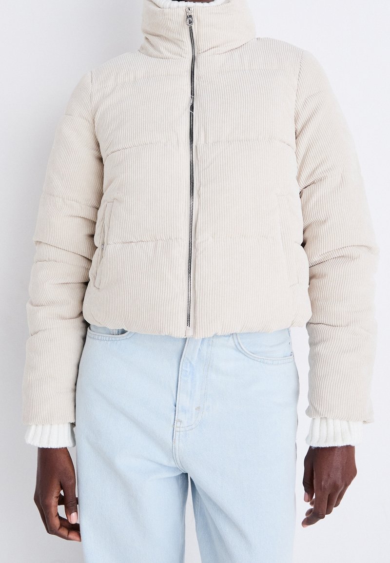 Personne portant une doudoune côtelée beige avec une fermeture éclair à l'avant et un jean taille haute bleu clair, debout sur un fond blanc.