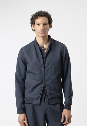 Joven con cabello rizado que lleva una chaqueta azul oscuro con cremallera y pantalones a juego, de pie frente a un fondo claro y liso.