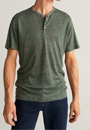 Man in een groen gemêleerd kortiemouwen overhemd met knopen en een donkere spijkerbroek, staand met zijn armen ontspannen langs zijn zij.