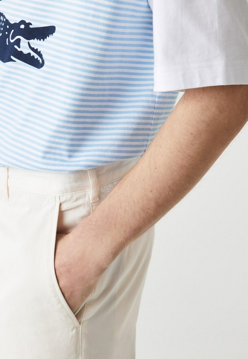 Persona che indossa pantaloni beige chiaro con una mano in tasca, abbinati a una camicia a righe bianco e azzurro chiaro caratterizzata da una grafica di un coccodrillo blu scuro.