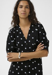 Fekete ing nagy fehér pöttyökkel, patentos elülső résszel, V-nyakkivágással és rövid ujjal. Ezüst hullámos karkötő a csuklón.