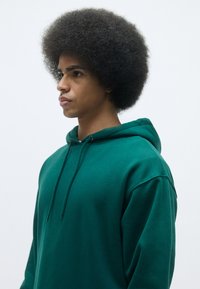 Felpa verde realizzata in tessuto testurizzato, con cappuccio regolabile e vestibilità comoda. Il modello si presenta di profilo, mettendo in evidenza il taglio di capelli.