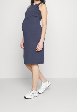 Gravid kvinde iført en ærmeløs knælængde navyblå kjole og hvide sneakers, stående mod en ensfarvet lys baggrund.
