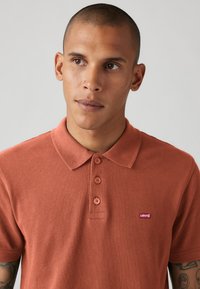 Cămașă polo de culoare ruginie, cu guler, trei butoane și un mic logo Levi's roșu pe partea stângă a pieptului. Fabricată dintr-un material texturat.