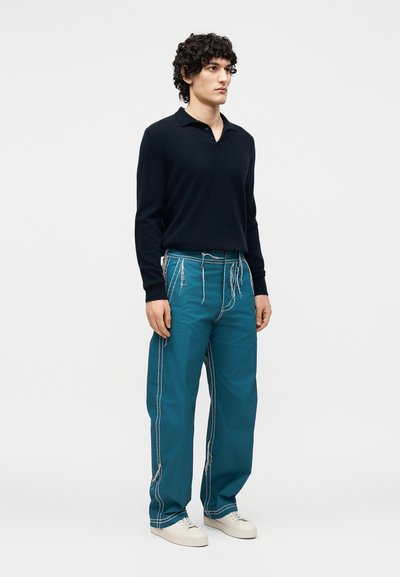 Νέος άνδρας στέκεται φορώντας μαύρο πόλο με μακριά μανίκια, τιρκουάζ παντελόνια με φαρδιά μπατζάκια και εμφανείς λευκές ραφές, και λευκά αθλητικά παπούτσια σε απλό φόντο.