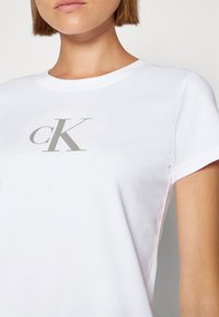 Biele bavlnené tričko s krátkym rukávom, s šedým logom "cK" potlačeným na textúrovanom štvorecovom pozadí. Minimalistický dizajn.